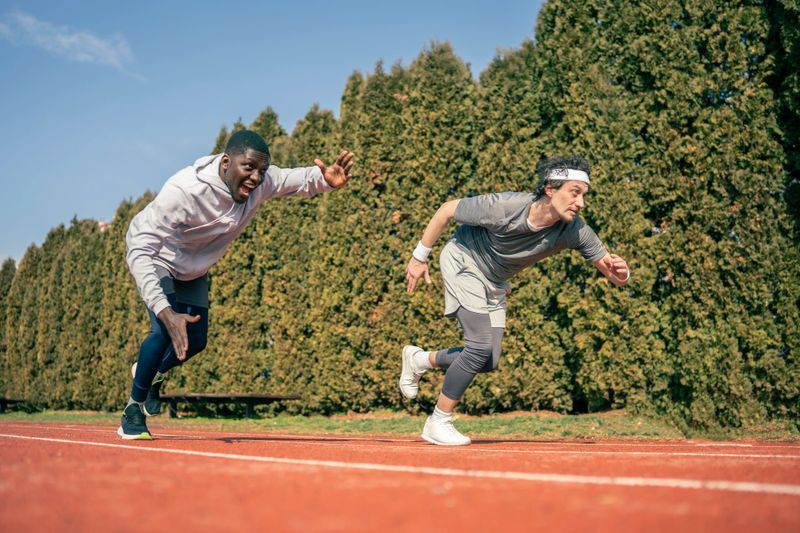 Two male athletes sprint on a red outdoor track during a training race, demonstrating speed, focus and competitive intensity under clear skies and warm sunlight.