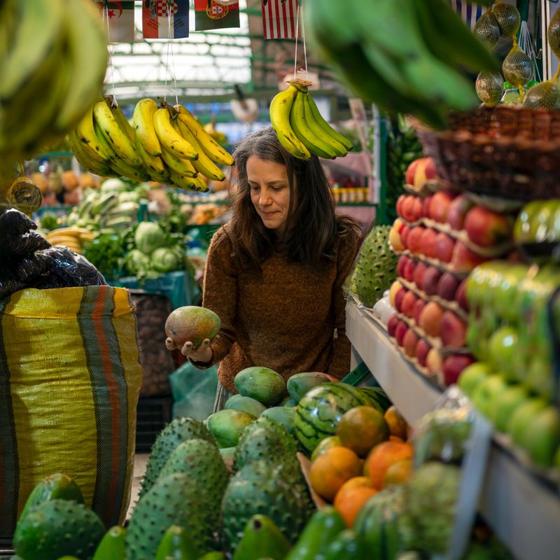 Adult woman selecting a mango at a colorful farmers market surrounded by fresh tropical fruits and vegetables. Lifestyle image about healthy eating, organic food shopping, and sustainable local markets.