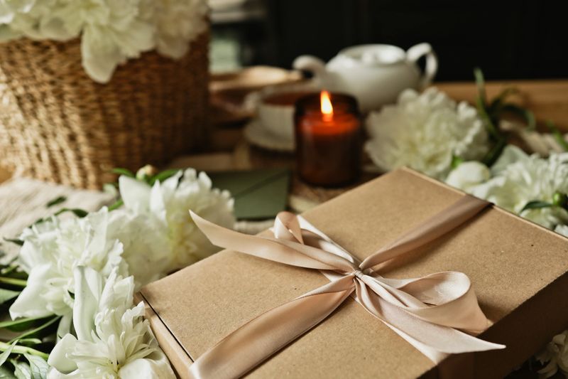A beautifully arranged scene featuring a gift box tied with a ribbon, surrounded by white peonies, a lit candle, and a teapot, evoking a sense of calm and relaxation