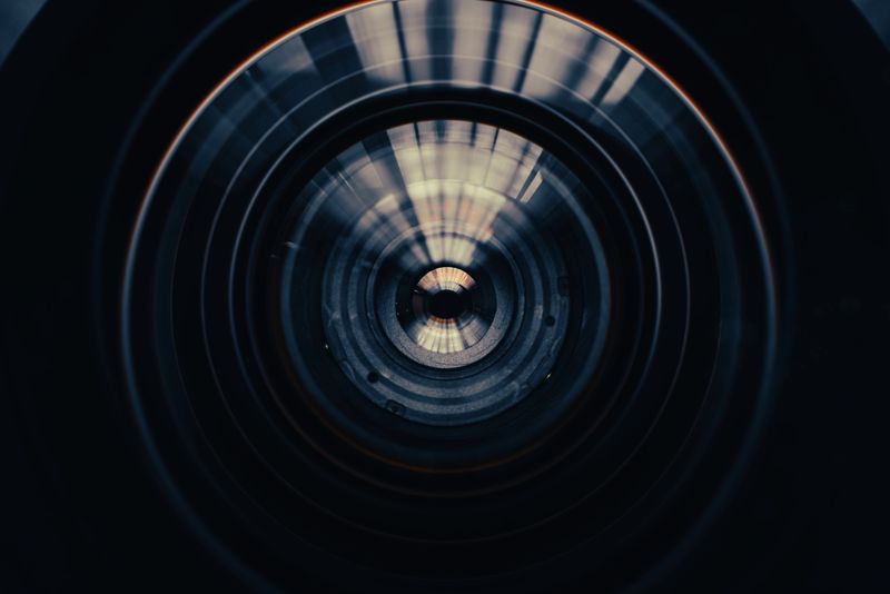 A macro shot looking directly into a professional camera lens, featuring complex concentric circles, dark metallic textures, and sharp light reflections creating a futuristic, high-tech, and abstract visual effect.