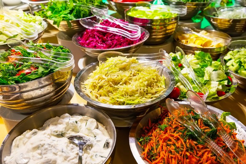 A colorful assortment of fresh vegetable salads, including tomatoes, cucumbers, carrots, and cabbage, are arranged in salad bowls on the buffet table.