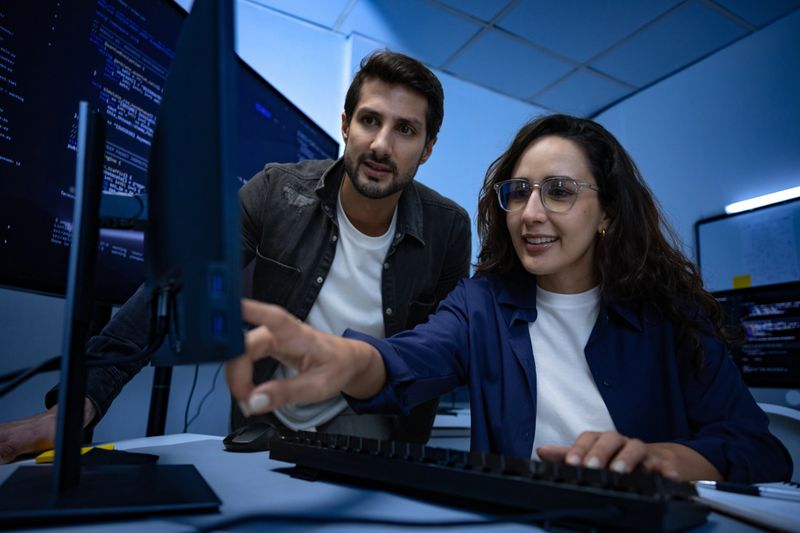 Focused male and female developer team collaborating on software code analysis in secure data center pointing at monitor screen with serious professional expressions solving complex cyber problem