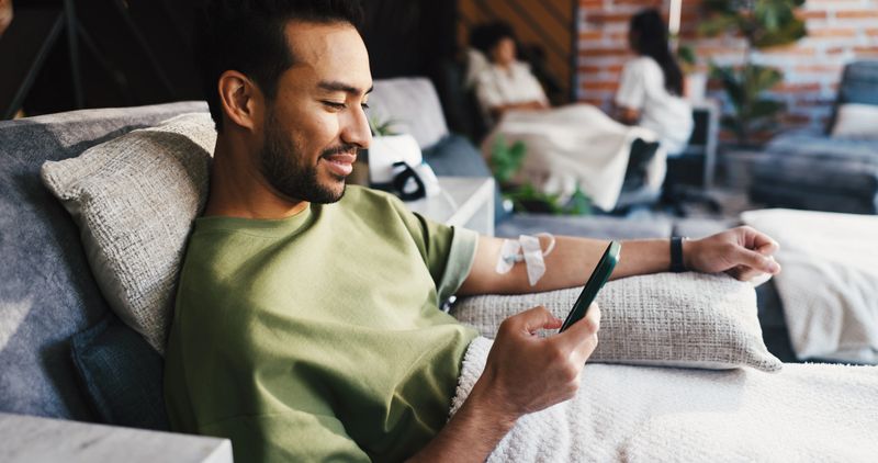 Phone, vitamin drip and man in lounge for immune system boost with social media or texting. Happy, iv detox and male person with cellphone for chatting on mobile app with benefits for wellness.