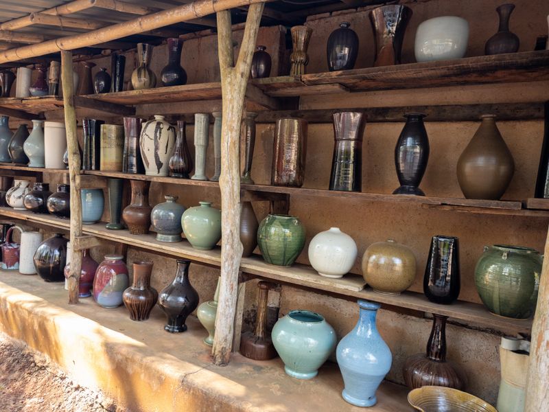 A vibrant collection of handcrafted ceramic vases and pottery pieces displayed on rustic wooden shelves in an artisanal outdoor market stall, showcasing diverse shapes and earthy glazes.