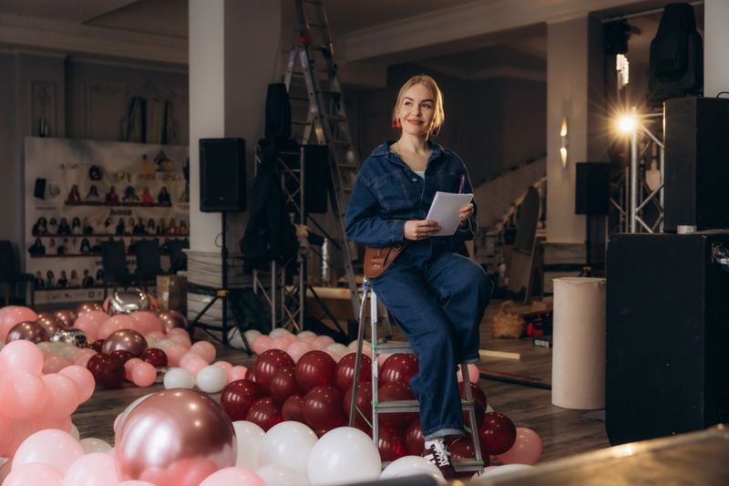 Woman event planner sitting on a ladder, smiling and holding notes, surrounded by balloons and equipment, decorating a celebration venue