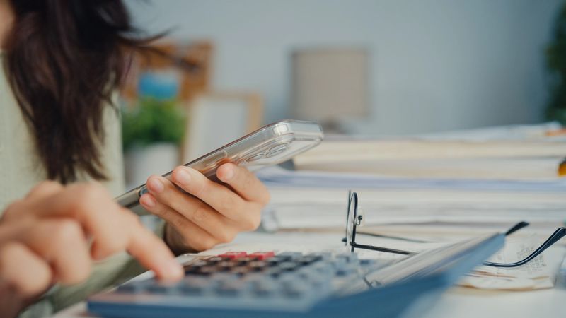Closeup of young woman checks expenses on smartphone while organizing bills and budgeting at home office. Personal finance concept.