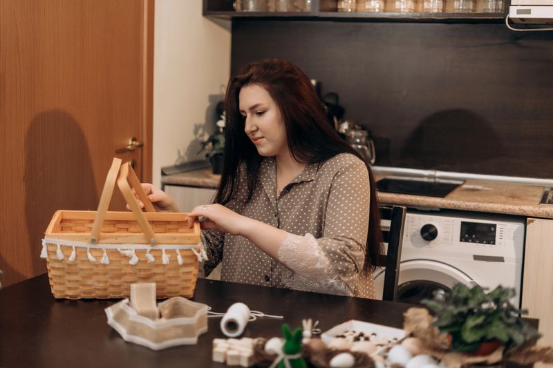 Woman at home in kitchen packing a wicker basket with gifts and decorations on table, cozy domestic lifestyle concept. Woman packing wicker gift basket in cozy home kitchen Woman packing wicker gift basket in cozy home kitchen. High quality photo