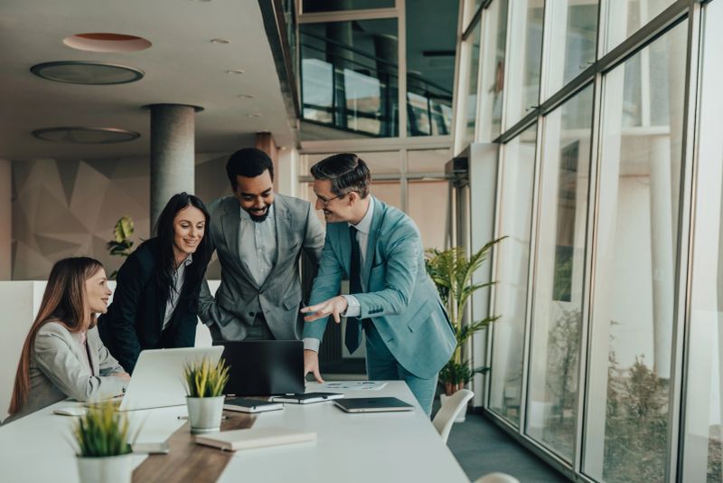 Business colleagues collaborate around a laptop in a modern office, smiling and discussing strategy, documents and ideas during a team meeting that conveys teamwork, professionalism and productivity.