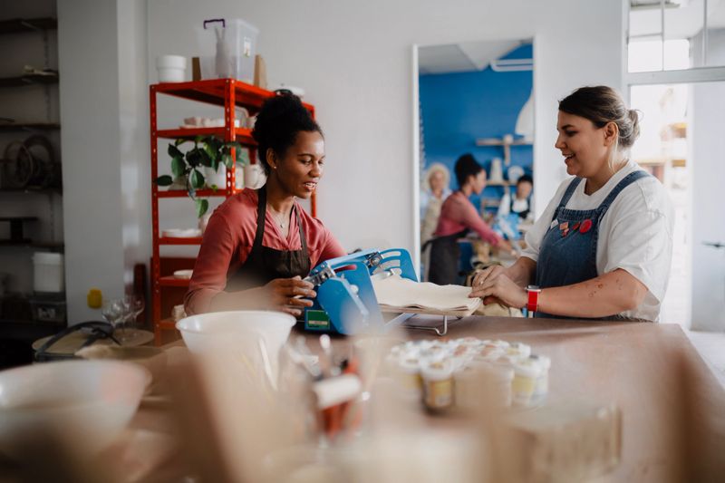 Two women collaborating in a pottery workshop, engaged in crafting and handling ceramic pieces. Surrounded by clay, tools, and handmade pottery, they are focused on their creative work in a warm and artistic environment. The image reflects craftsmanship, teamwork, creativity, and the spirit of small artisanal businesses.