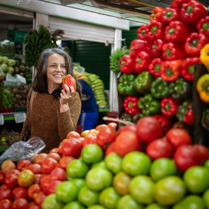 Mid adult woman smiling while selecting a fresh red bell pepper at a colorful outdoor farmers market. Vibrant display of tomatoes and green peppers in the foreground. Concept of healthy eating, grocery shopping, organic food and everyday lifestyle.