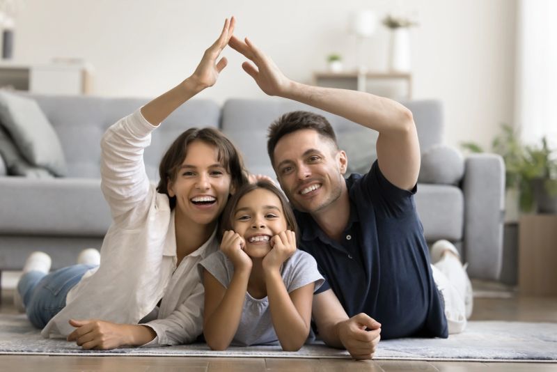 35s Latin couple and little adorable daughter lying on warm floor, making roof symbol with joined hands, smiling, look at camera. Real estate, house buying, insurance, bank mortgage for young family