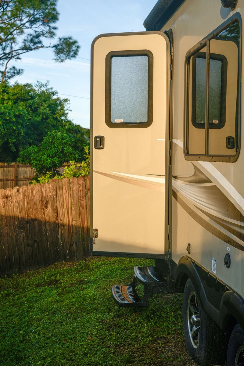 Vertical photo of open doors on a beige RV trailer, parked on green grass in a forest, with sunlight streaming through, a cozy and inviting outdoor atmosphere