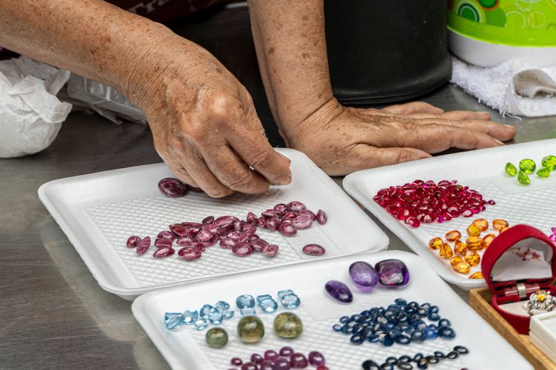 Close-up of a gem trader carefully sorting and grading polished gemstones arranged on white trays. Various colorful precious and semi-precious stones prepared for evaluation, trading, and jewelry manufacturing.