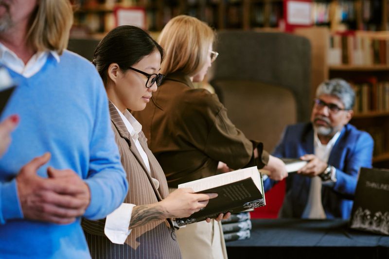 Asian young adult woman reading book while standing in line at book presentation event, Caucasian young adult woman receiving signed copy from middle aged South Asian man seated at table