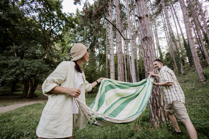 Couple setting up a hammock while preparing for a camping trip in nature. Man and woman enjoying outdoor adventure, teamwork, and relaxation before settling into their campsite. Concept of camping lifestyle, travel, leisure time, and connection with nature.
