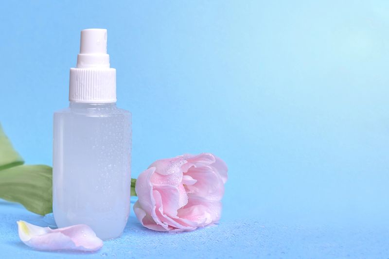 Clear spray bottle positioned next to a pink flower and petals on a soft blue background, creating a fresh and clean aesthetic