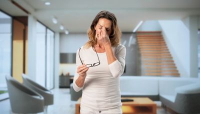 A woman in white rubbing her eyes and holding glasses indoors.
