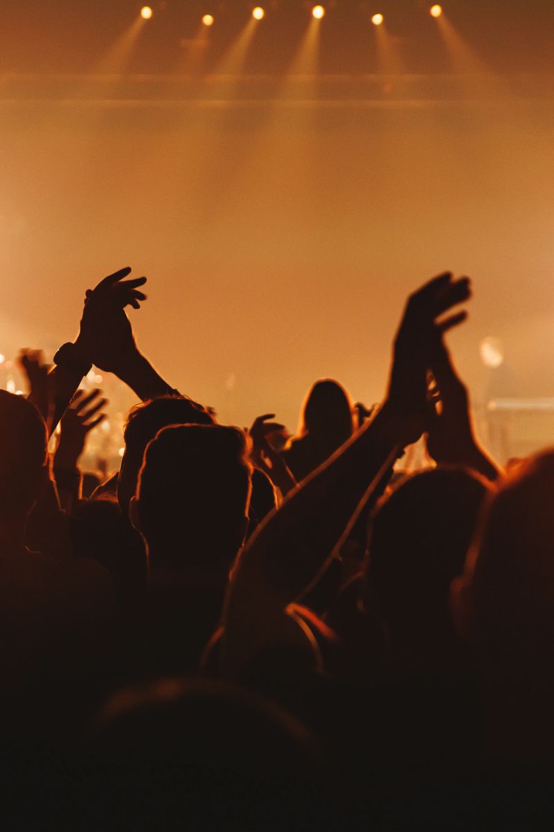 View from the crowd showing people with their hands raised and clapping in rhythm with the music. Warm stage lighting illuminates the haze and creates a vibrant atmosphere.