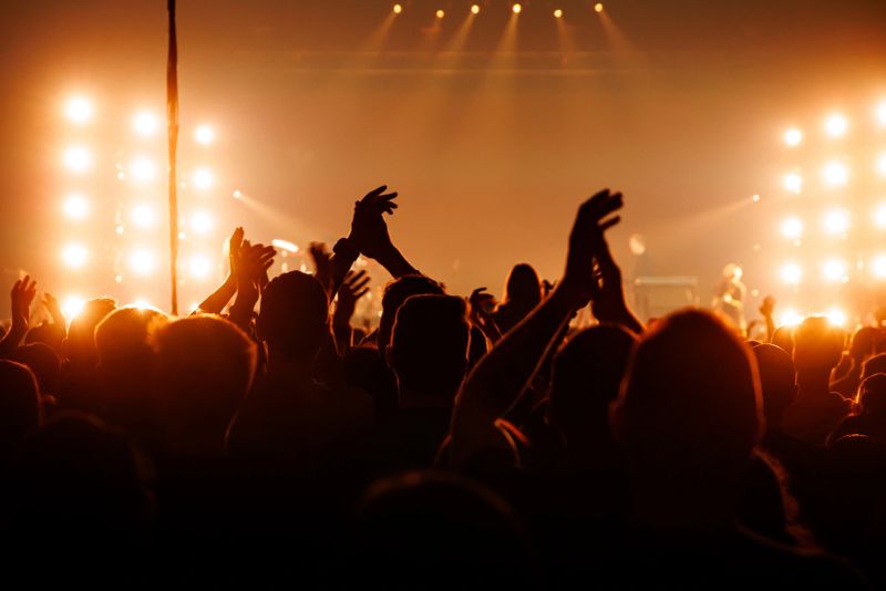 An enthusiastic audience applauds a performer during a live show. Bright orange stage lights create a high-contrast silhouette of the cheering fans.