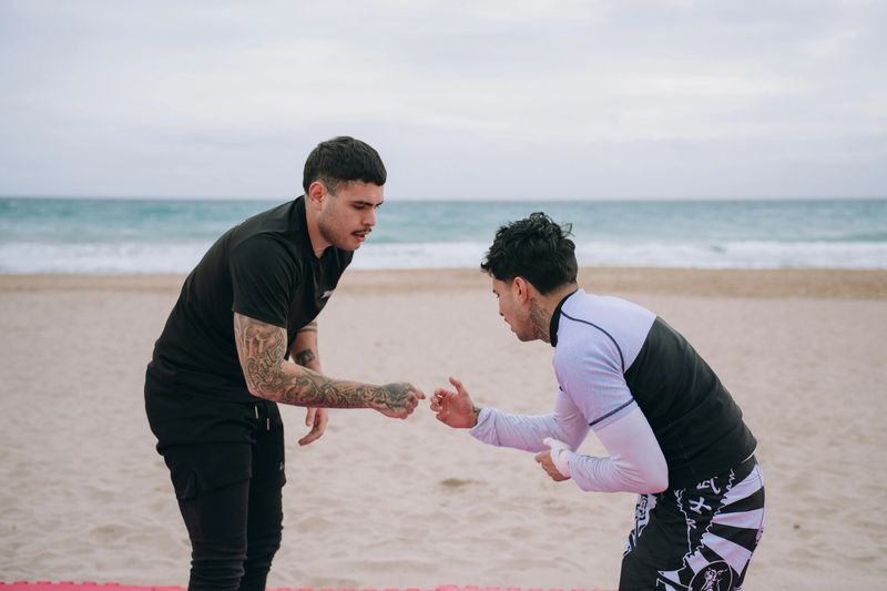 Two young adult men training mixed martial arts on beach, practicing grappling techniques on a mat by the ocean