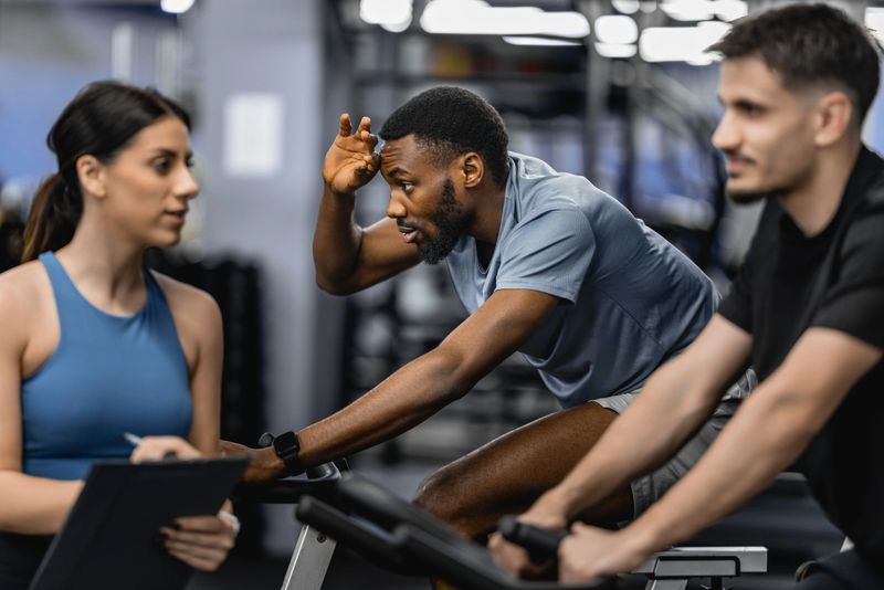 Three adults participate in an intense indoor cycling session as a trainer coaches technique and motivation, conveying energy, focus, teamwork and determination during a modern gym cardio workout.