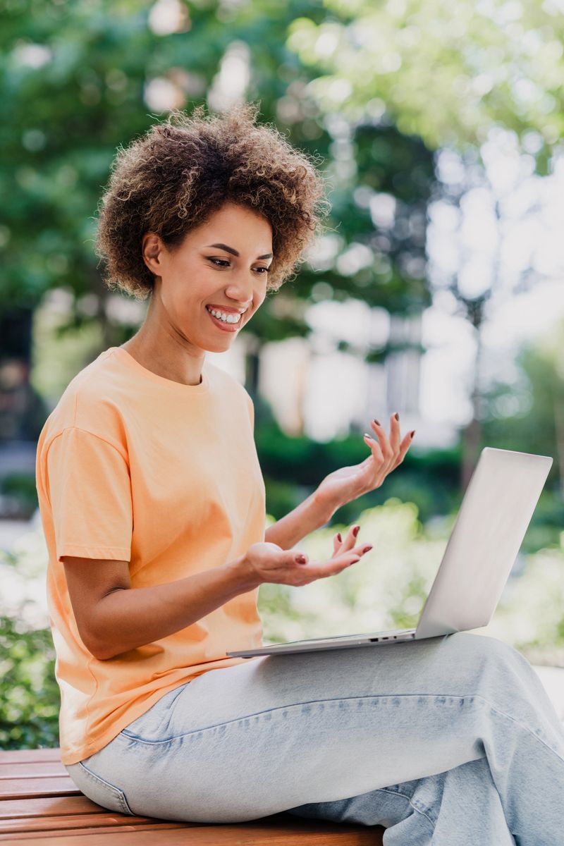 A joyful African American woman in a peach Tshirt and jeans sits on a wooden bench outdoors, using a laptop, blending work and leisure in a bright city park on a sunny day.