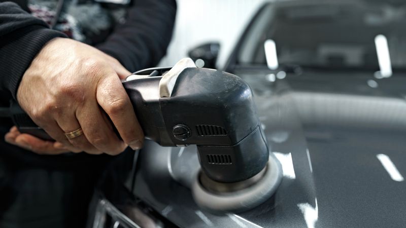 A person holds a polisher as they work on a car's surface in a workshop setting.