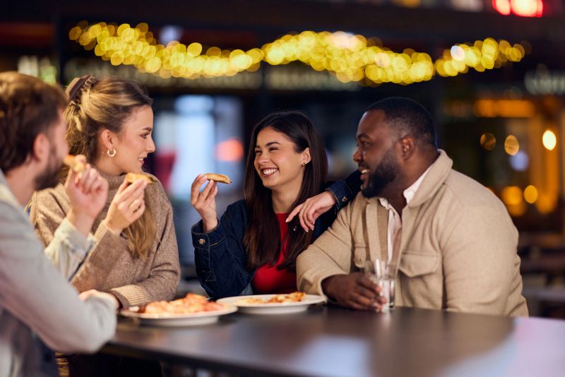 A diverse group of friends shares pizza and conversation at an evening outdoor setting, illuminated by warm string lights. African-American man and friends laugh, strengthening bonds, suitable for lifestyle, friendship, and casual dining campaigns.