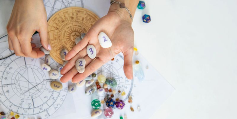 Astrologist hands holding numerology stones over natal charts and zodiac wheel.