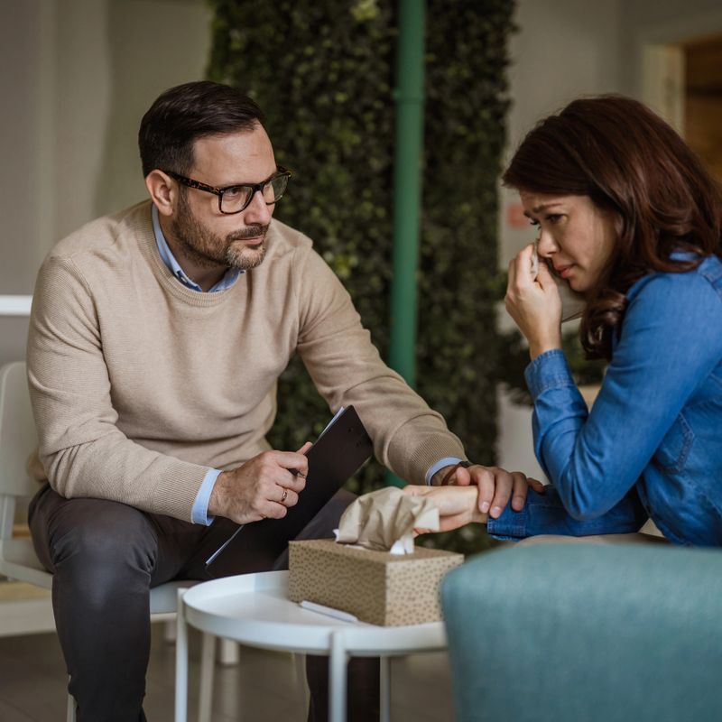 Male psychologist listening intently to a distressed young woman covering her face with a tissue, offering support and guidance in a private counseling session about mental health