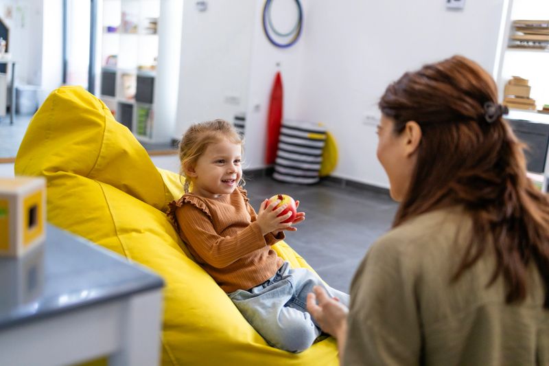 Young girl sits on a yellow lazy bag, smiling and holding an apple while attentively listening to a female child psychologist during a bright, friendly therapy session.