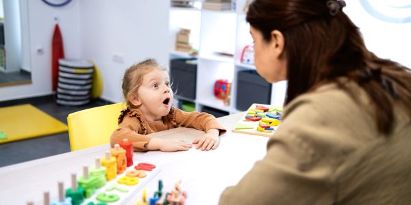 A surprised child interacting with an adult at a table in a classroom.