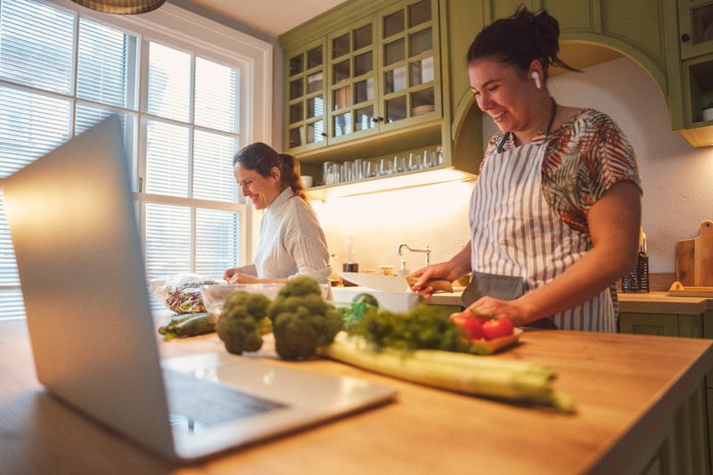 Smiling Asian woman making detox salad in home kitchen with friend, watching recipe on laptop. Gut health, microbiome, nutritionist podcast, mindful eating & active life balance
