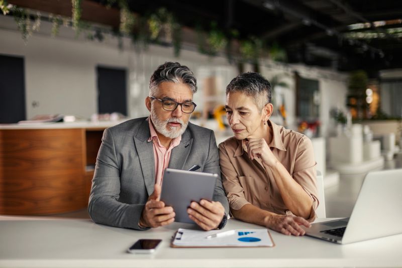 Two mature adult business professionals are collaborating, looking at a digital tablet and discussing data while working at a modern office desk, showing teamwork and shared innovation