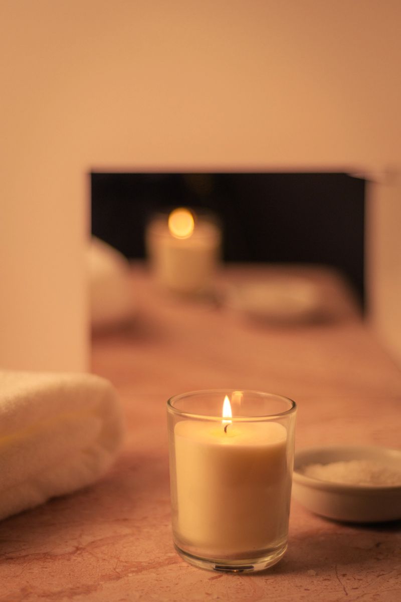 Vertical shot of lit candle with mirror bokeh, towel and bowl of bath salts on counter. Minimal cozy home spa atmosphere, warm tones and copy space.