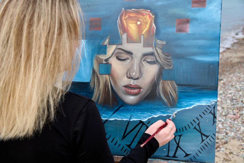 Young woman artist with blonde hair paints surreal portrait featuring ocean waves and fragmented face on canvas outdoors, capturing creativity and focus