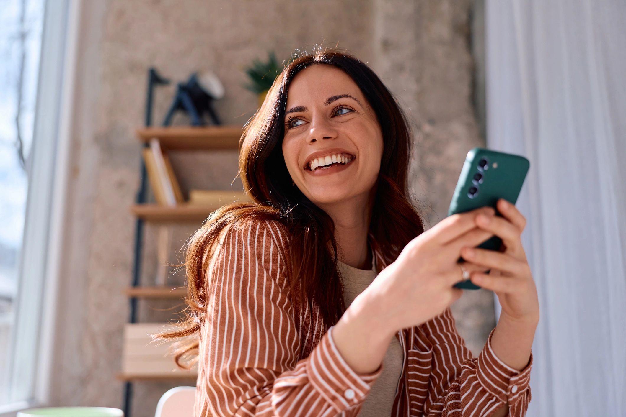 Woman in a striped shirt sitting near a window and using a smartphone, looking away cheerfully while laughing, expressing concepts of joy, communication, technology, and connection