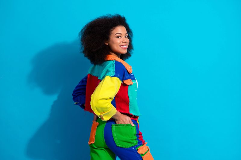 A stylish young woman wearing a multicolor patched jacket and jeans enjoys a cheerful moment against a vibrant blue backdrop. The image conveys fashion diversity confidence and playful energy suitable for lifestyle and promotional use.