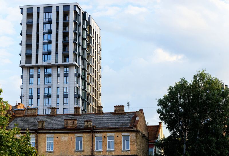 A contemporary residential tower stands tall behind weathered, historic brick buildings, set against a backdrop of soft, diffused clouds