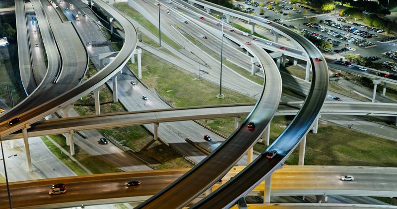 Aerial photograph of a multi-level highway interchange in Dallas, Texas, captured during the day. The complex infrastructure features sweeping curved ramps and overpasses connecting multiple freeways, with vehicles traveling along the various roadways. The interchange demonstrates the extensive transportation network that serves the Dallas metropolitan area, with concrete support pillars and barriers defining the different traffic levels. Parking areas and green spaces are visible adjacent to the highway system, illustrating the urban planning that accommodates both vehicular infrastructure and surrounding development in this major Texas city.   • Authorization was obtained from the FAA for this operation in restricted airspace.
