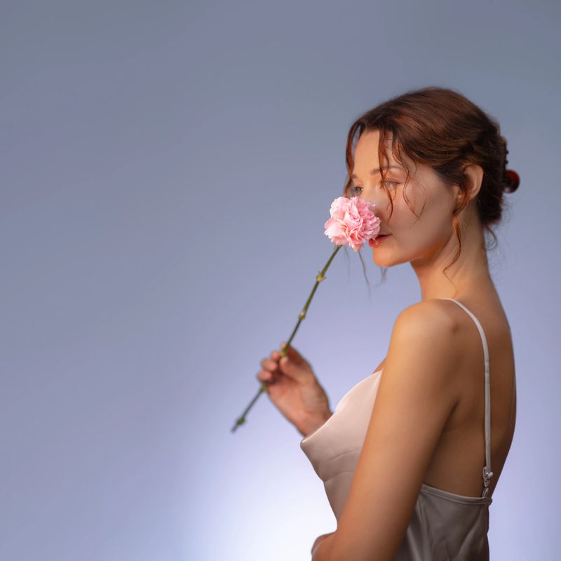 Graceful woman in soft lighting poses with a delicate pink carnation, expressing femininity and elegance. Perfect for romantic, beauty, skincare, and floral advertisement or editorial use.