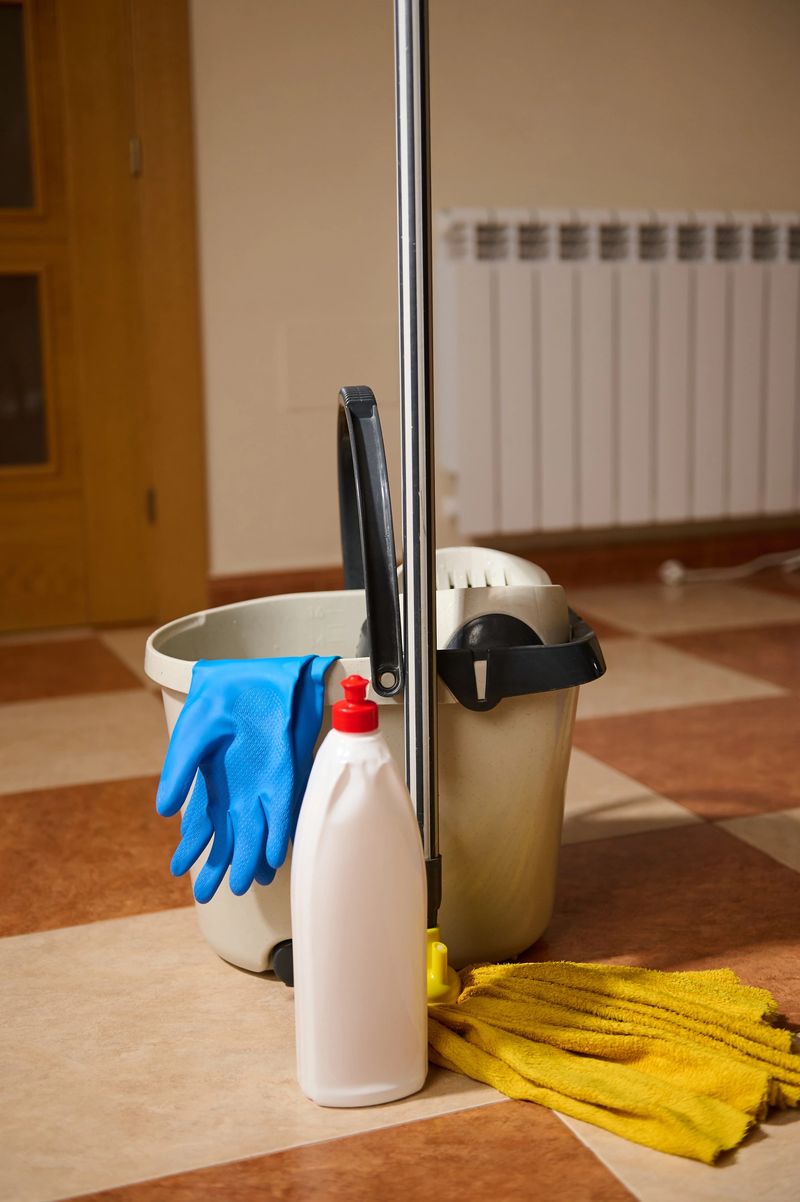 Middle-aged woman cleaning a tiled home floor with mop, bucket, rubber gloves and detergent, portraying domestic chores, household care and everyday home maintenance.