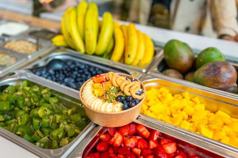Acai bowl topped with bananas, blueberries, mango, kiwi, strawberries, and peanut butter is surrounded by stainless steel trays filled with various fresh fruits for a healthy snack or breakfast