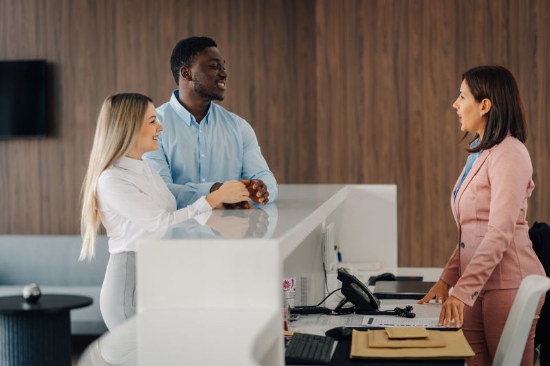 Diverse smiling couple interacts with friendly hotel front-desk agent during check-in at a modern, elegant lobby, welcoming service
