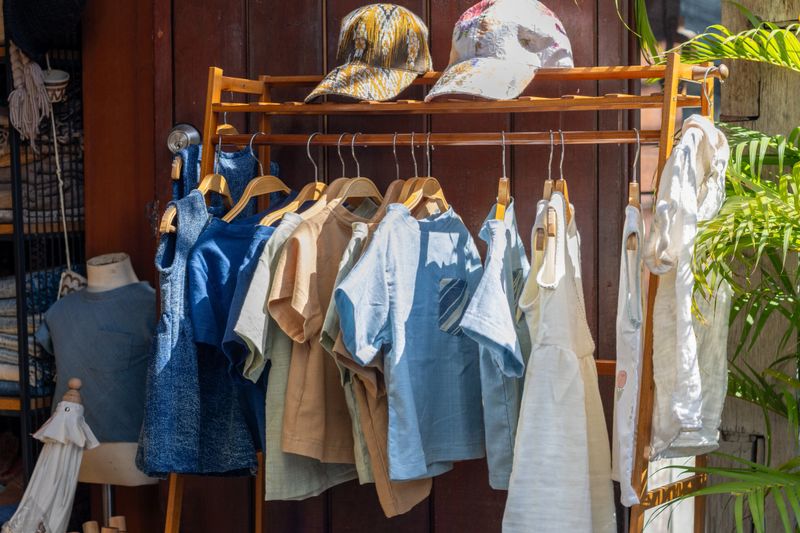 A wooden clothes rack showcases a collection of natural fabric garments like dresses, shirts, and tops in varying blue, earth tones, and white. This outdoor boutique display highlights sustainable fashion and handmade apparel in a relaxed setting.