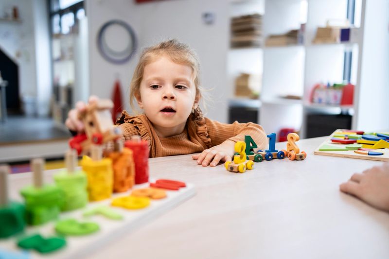 Preschool-aged child engaging in educational play. Developing cognitive skills and fine motor abilities by sorting colorful number and shape toy elements on a light table in a bright classroom setting.