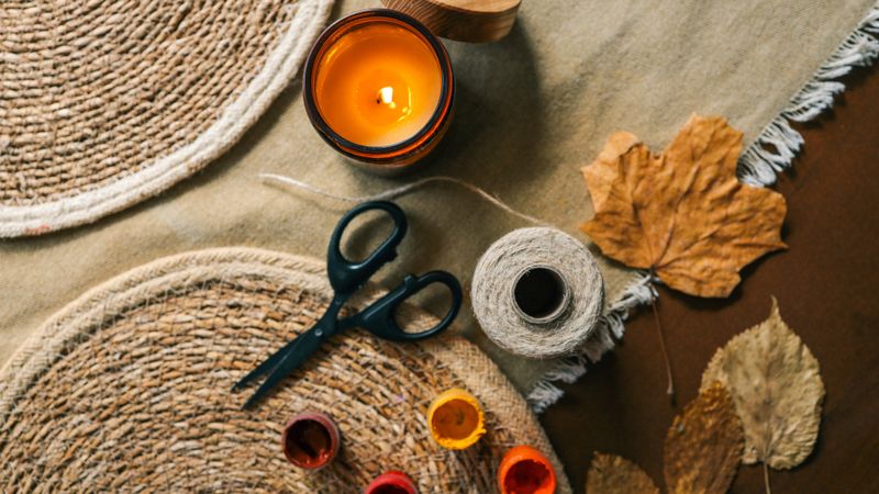 A top-down view of crafting materials including scissors, twine, paints, a lit candle, and fallen leaves arranged on a rustic textile surface