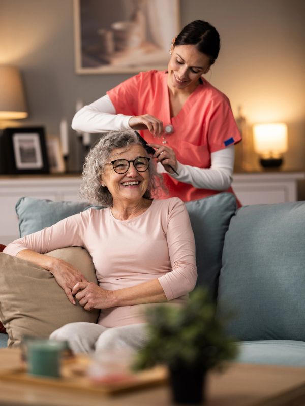 Caregiver gently combing elderly woman's hair, both smiling in a cozy living room.