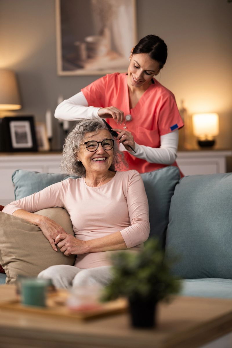 Young cheerful nurse combing senior woman's hair while she is sitting on couch in living room, talking and smiling.