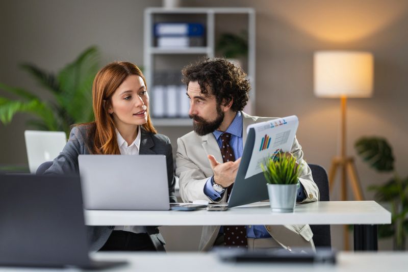 Two business colleagues in an office discussing charts and financial reports while collaborating on strategy, using laptops and printed graphs in a modern professional workspace.